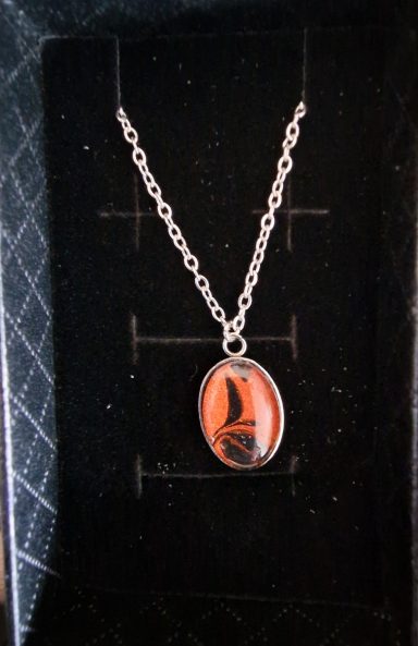 Stainless steel oval pendant containing parts of original art orange and black with gold shimmer on a black background.
