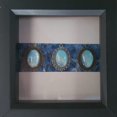 3 oval pendants containing light blue and silver artwork resting on dark blue rectangle on white background in black frame